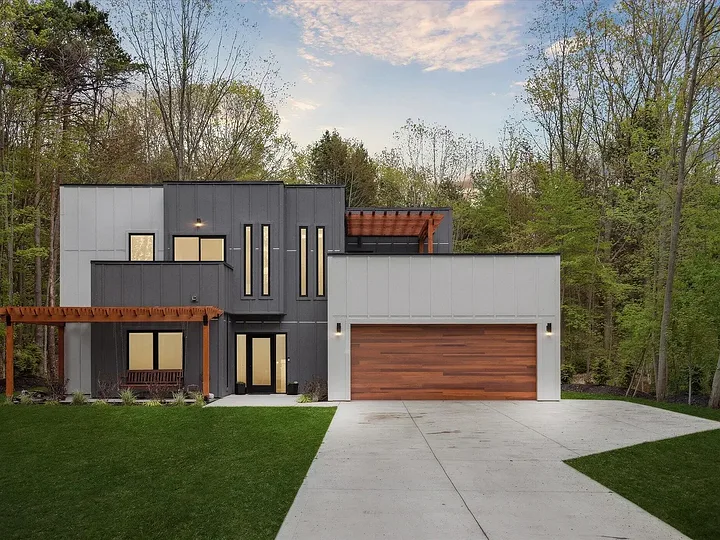 Modern two-story home with white and charcoal panel siding, a cedar garage door, and cedar pergolas on a wooded lot