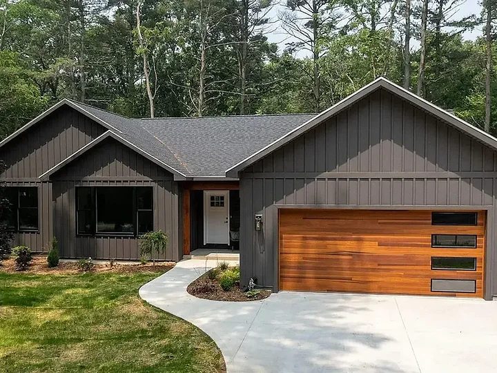 Dark board-and-batten modern farmhouse with a cedar garage door, set among tall pines